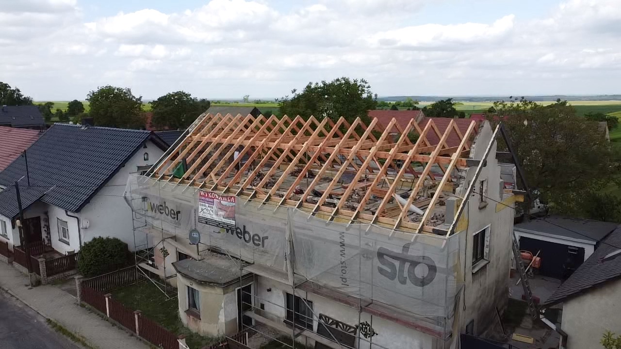 Dom jednorodzinny w trakcie wymiany konstrukcji dachu, widoczne drewniane krokwie i łaty, rusztowania z siatką ochronną z reklamą Weber i Sto, w tle inne domy i pola uprawne.