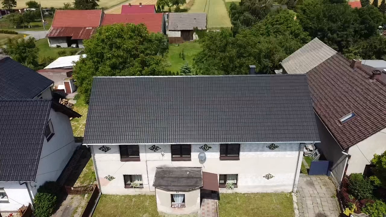 Widok z góry na dom z ciemnoszarą dachówką ceramiczną, białą elewacją i dekoracyjnymi elementami pod oknami, otoczony zielenią i innymi budynkami w oddali.