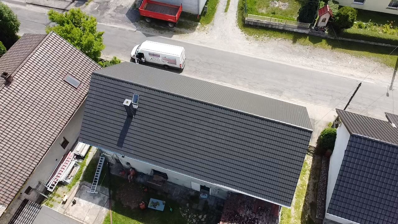Widok z góry na dom z ciemnoszarą dachówką, białą obudową komina i nowo wykończonym dachem; w tle biały van firmy budowlanej i czerwona przyczepa.