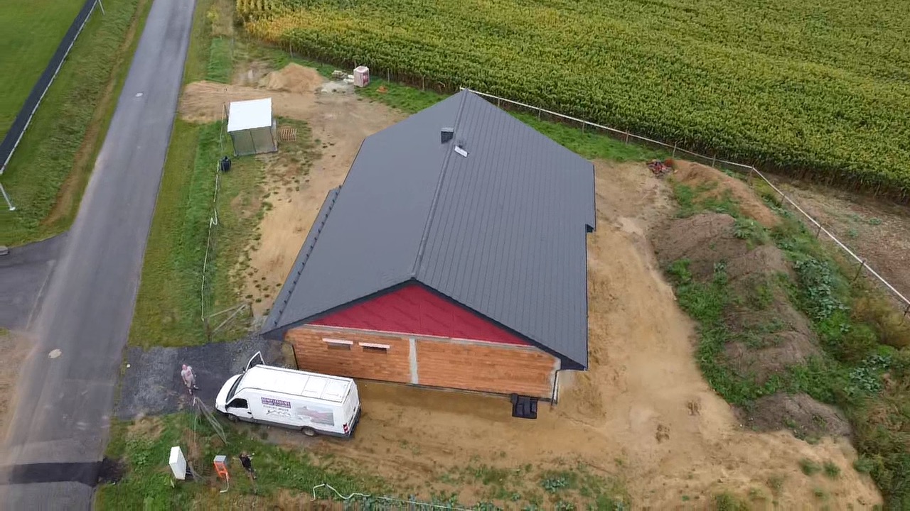 Widok z lotu ptaka na budynek w trakcie budowy z szarym dachem z blachy, czerwonymi elementami pod dachem i częściowo wykończonymi ścianami z cegły, obok stoi biały samochód dostawczy.