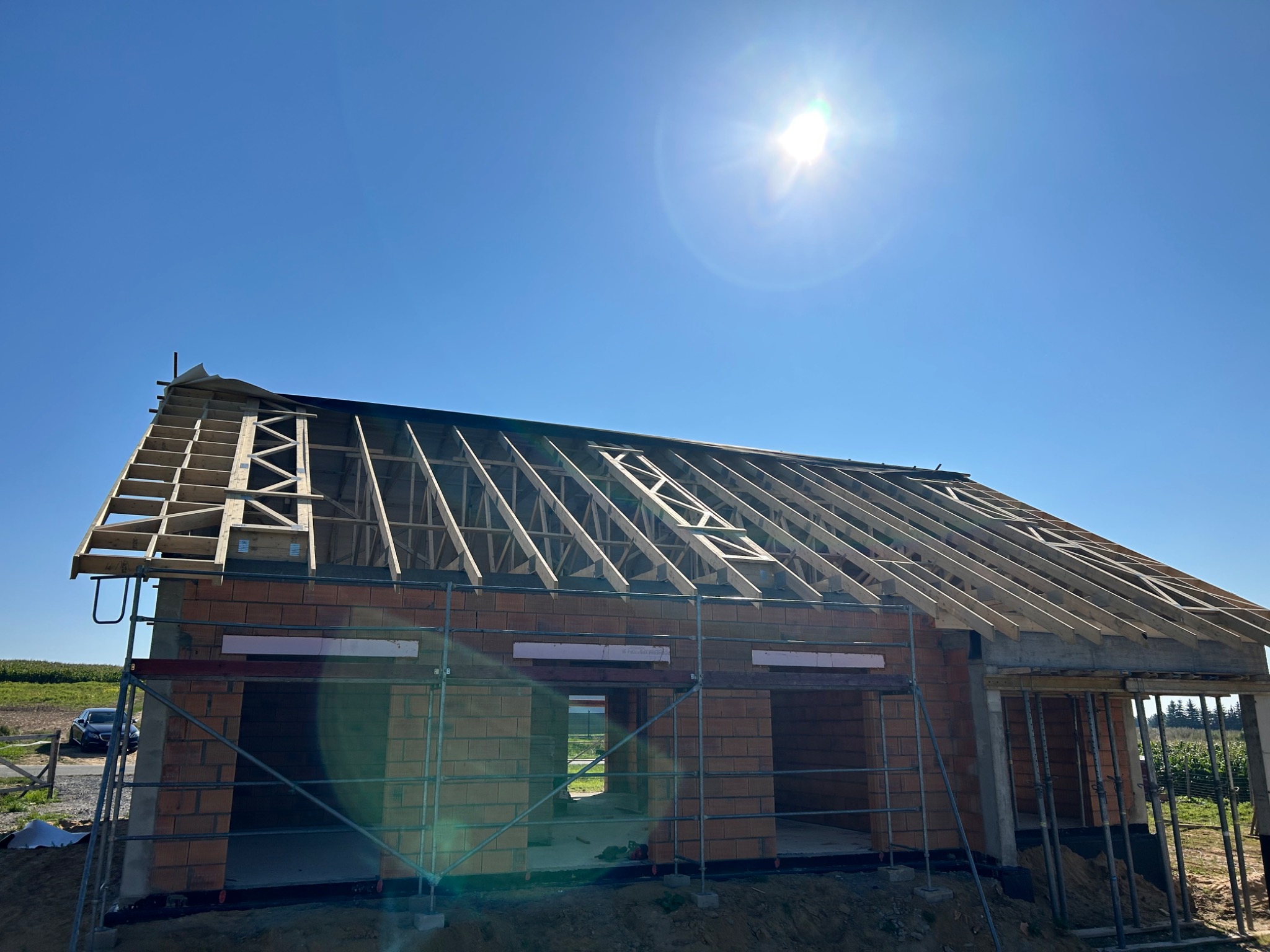 Budowa domu jednorodzinnego w stanie surowym otwartym, widoczna więźba dachowa z deskowaniem, murowane ściany z czerwonej cegły, rusztowania, błękitne niebo i ostre słońce.