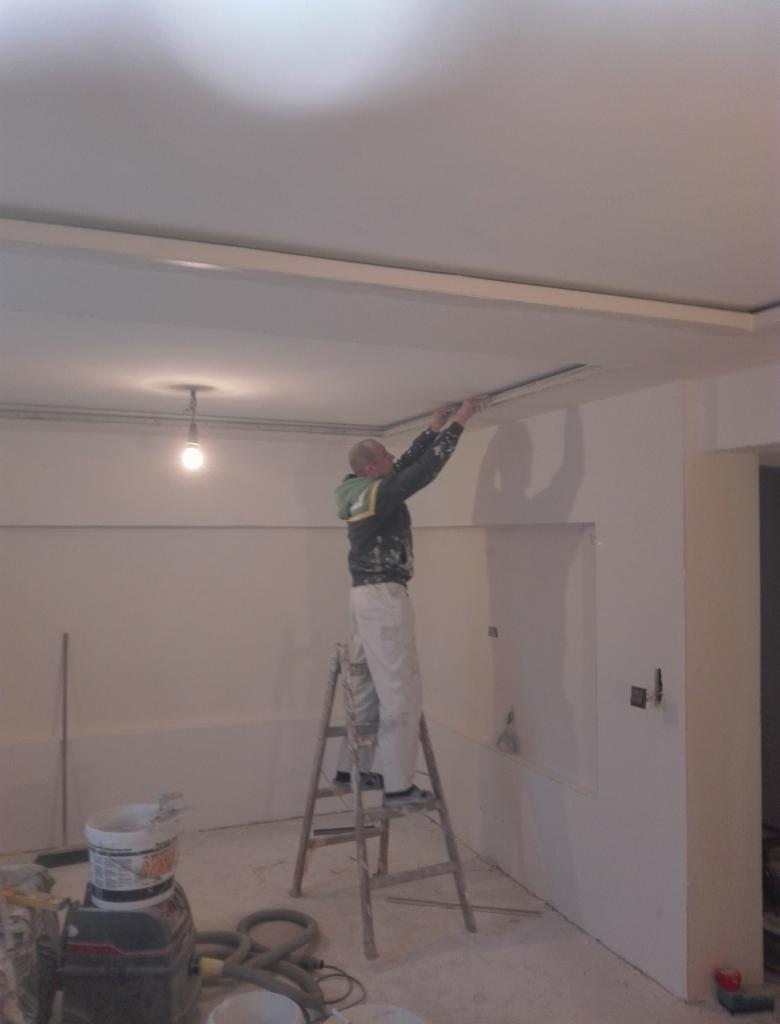 Pracownik na drabinie montuje listwę oświetleniową pod sufitem podwieszanym, widoczny sprzęt malarski i odkurzacz przemysłowy.