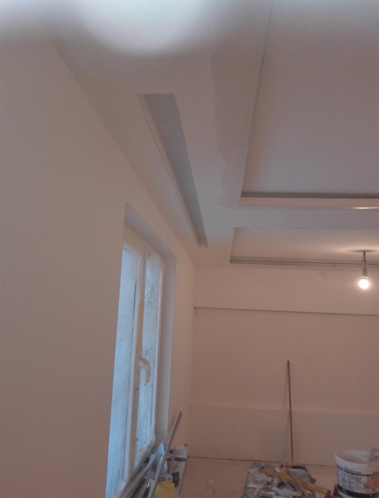 Wnętrze w trakcie prac wykończeniowych: świeżo pomalowane ściany w jasnym odcieniu, widoczne elementy sufitu podwieszanego z oświetleniem, okno zabezpieczone folią, na podłodze narzędzia i puszki...