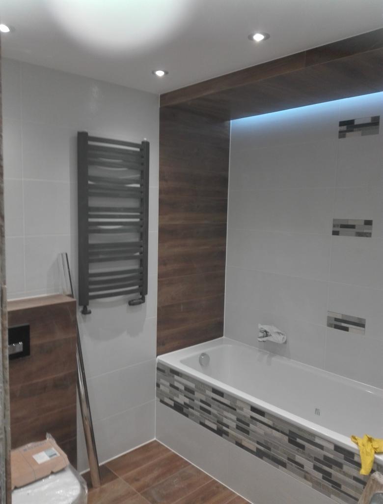 Nowoczesna łazienka z wanną obudowaną mozaiką, grzejnikiem drabinkowym na ścianie i drewnopodobnymi panelami na ścianie oraz suficie z oświetleniem punktowym.