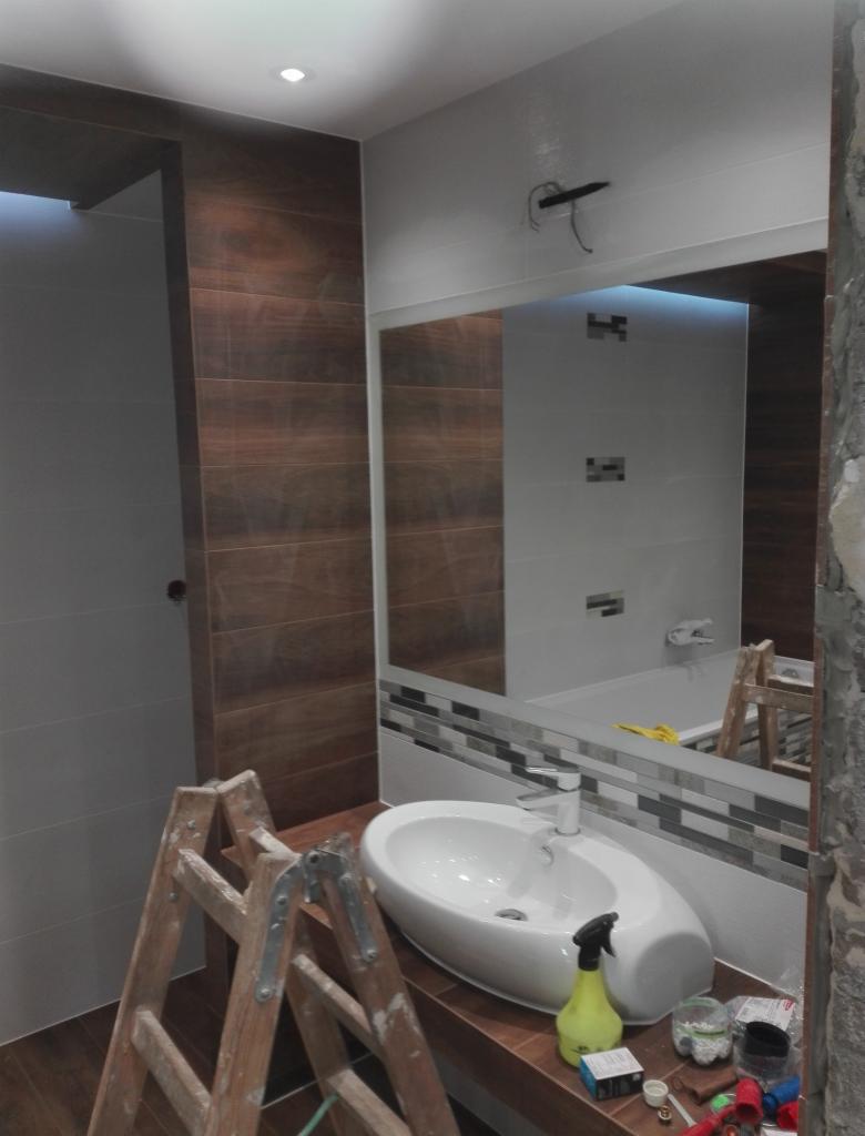 Łazienka w trakcie prac: drewnopodobne płytki ścienne, biała umywalka na brązowym blacie, drabina malarska, widoczne narzędzia i chemia budowlana.