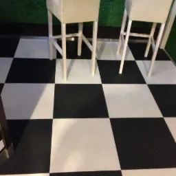 Czarno-biała podłoga w szachownicę z płytek samozamykających się R-Tile, widoczne dwa białe krzesła barowe i fragment zielonej ściany.