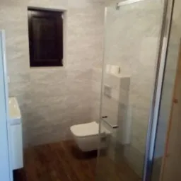 Nowoczesna łazienka z drewnianą podłogą, ścianami wykończonymi płytami imitującymi marmur, kabiną prysznicową ze szkła hartowanego i podwieszaną toaletą.