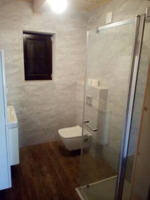 Nowoczesna łazienka z drewnianą podłogą, ścianami wykończonymi płytami imitującymi marmur, kabiną prysznicową ze szkła hartowanego i podwieszaną toaletą.