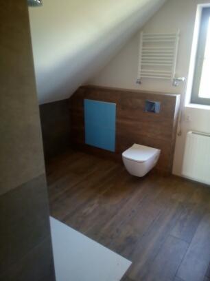 Nowoczesna łazienka na poddaszu z drewnopodobną podłogą, wiszącą toaletą i zabudową z akcentem w kolorze błękitnym, widoczny grzejnik drabinkowy i okno.