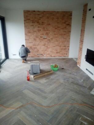 Pracownik w trakcie układania cegieł dekoracyjnych na ścianie w salonie z parkietem w jodełkę; widoczne narzędzia, pojemnik z gąbkami i butelka napoju.