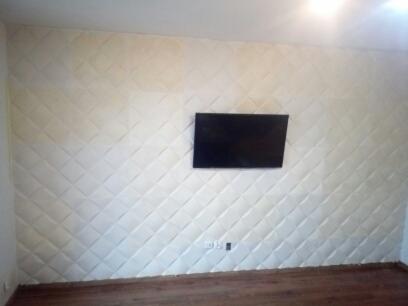 Ściana wykończona panelami tapicerowanymi w romby, z zamontowanym telewizorem, widoczne gniazdka elektryczne i fragment drewnianej podłogi.