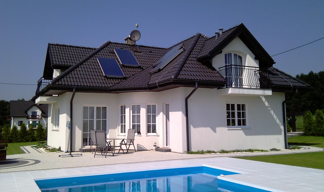 Nowoczesny dom jednorodzinny z basenem, panelami słonecznymi na dachu i balkonem z metalową balustradą. Biała elewacja, ciemna dachówka i starannie zagospodarowany ogród.