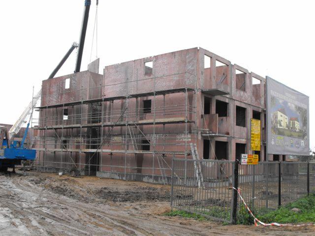 Montaż prefabrykowanego budynku wielorodzinnego z rusztowaniami i dźwigiem, widoczny baner reklamowy inwestycji. Błotniste podłoże.