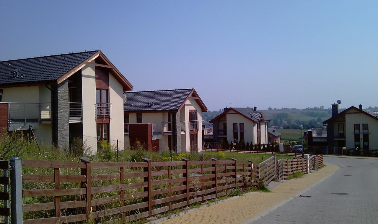 Osiedle domów jednorodzinnych z drewnianym ogrodzeniem, widok z ulicy. Domy posiadają dwuspadowe dachy z ciemnej dachówki i elementy dekoracyjne z kamienia.