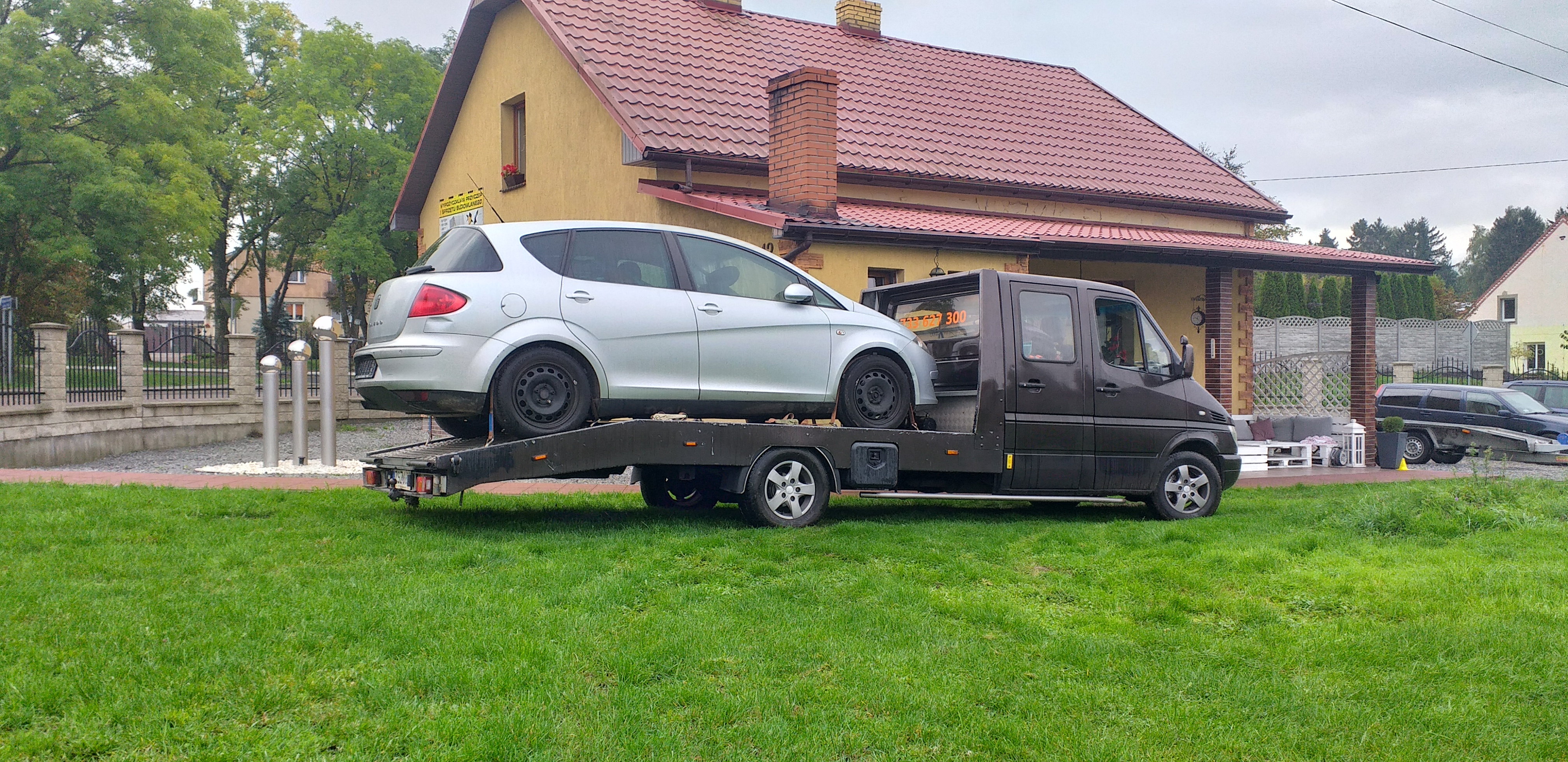Laweta z srebrnym samochodem osobowym Seat Toledo na platformie, zaparkowana na trawniku przed budynkiem z czerwoną dachówką.