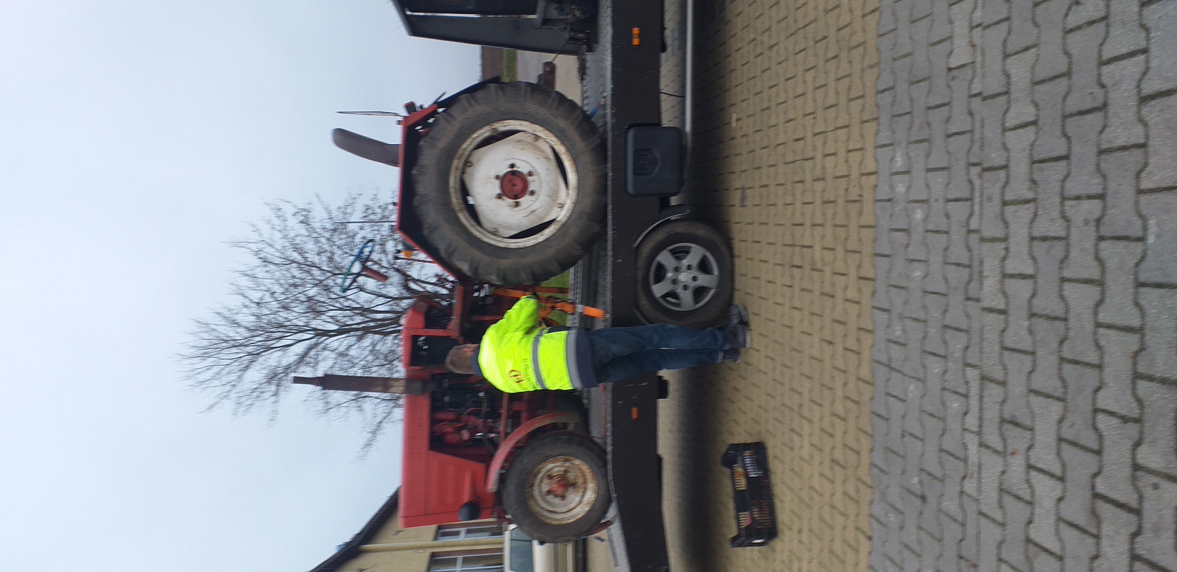 Czerwony traktor umieszczony na platformie transportowej, zabezpieczony pasami, pracownik w żółtej kamizelce kontroluje mocowanie, w tle widoczne drzewo bez liści.