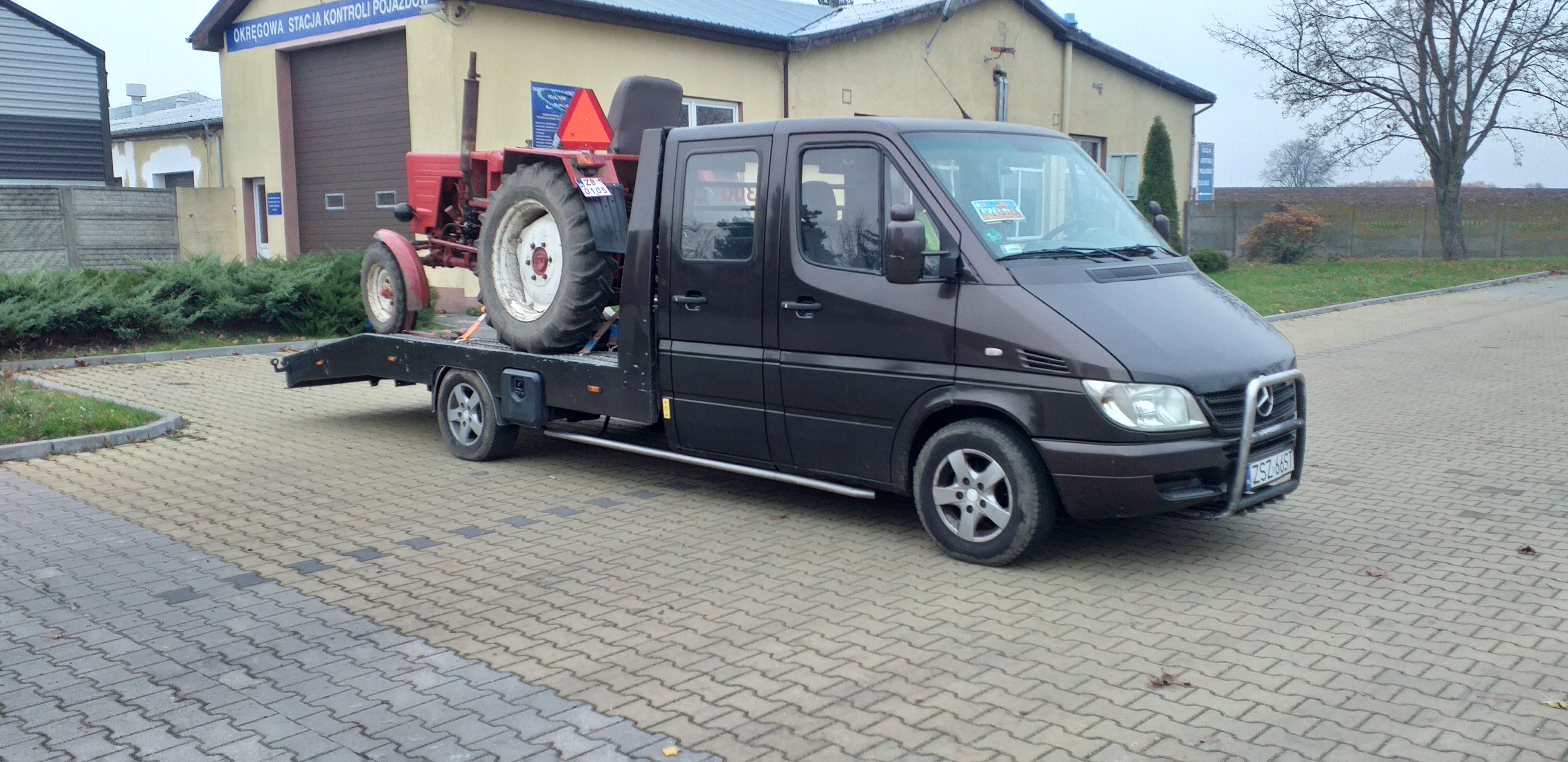 Brązowy van z platformą transportową przewozi czerwony traktor rolniczy na tle budynku Okręgowej Stacji Kontroli Pojazdów.