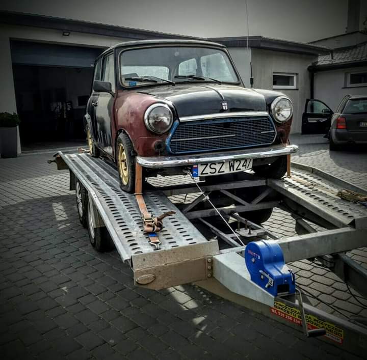 Czarno-czerwony, zabytkowy Mini Cooper z widoczną rdzą, transportowany na metalowej lawecie z niebieską wciągarką, zaparkowanej na brukowanym podjeździe przed garażem.