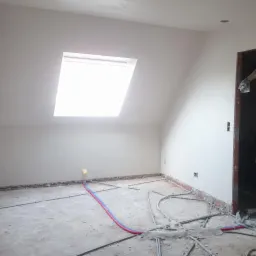 Poddasze po nałożeniu gładzi gipsowej, widoczne rury instalacji grzewczej na podłodze, okno dachowe w skośnej ścianie i fragment murarza podczas pracy w przejściu.