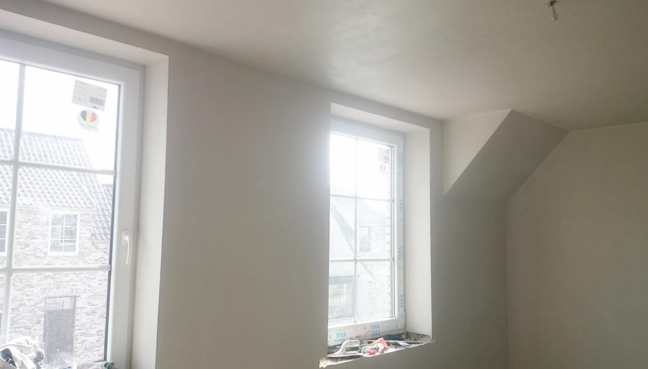 Świeżo położone, jasne wykończenie ścian w pokoju z dwoma oknami, widoczne narzędzia i resztki materiałów budowlanych na parapecie okiennym.