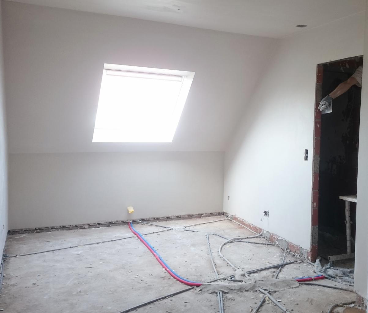 Poddasze po nałożeniu gładzi gipsowej, widoczne rury instalacji grzewczej na podłodze, okno dachowe w skośnej ścianie i fragment murarza podczas pracy w przejściu.