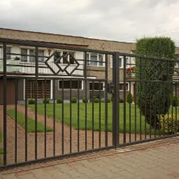 Otwarta, metalowa brama wjazdowa w kolorze grafitowym, z ozdobnymi rombami, prowadząca do domu z zielonym trawnikiem i ścieżką z kostki.