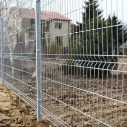 Nowoczesne, ocynkowane ogrodzenie z paneli stalowych na tle domu jednorodzinnego i drzew, widoczne mocowania słupków.