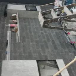 Wnętrze w trakcie prac wykończeniowych: szare płytki imitujące cegłę na ścianie, białe płytki podłogowe, aluminiowe profile, drabina malarska i tuby z silikonem.