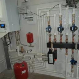 Węzeł cieplny z kotłem gazowym, pompami obiegowymi i zbiornikami wyrównawczymi w kotłowni wyłożonej białymi płytkami.