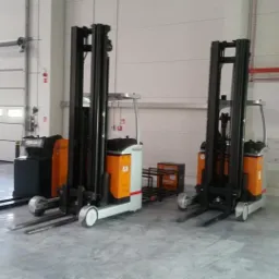 Dwa elektryczne wózki widłowe stojące w magazynie z widocznymi drzwiami segmentowymi i instalacją przeciwpożarową w tle.