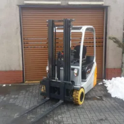 Elektryczny wózek widłowy marki Still, zaparkowany przed budynkiem z brązową bramą rolowaną i szyldem 'Forklift Truck Service'.