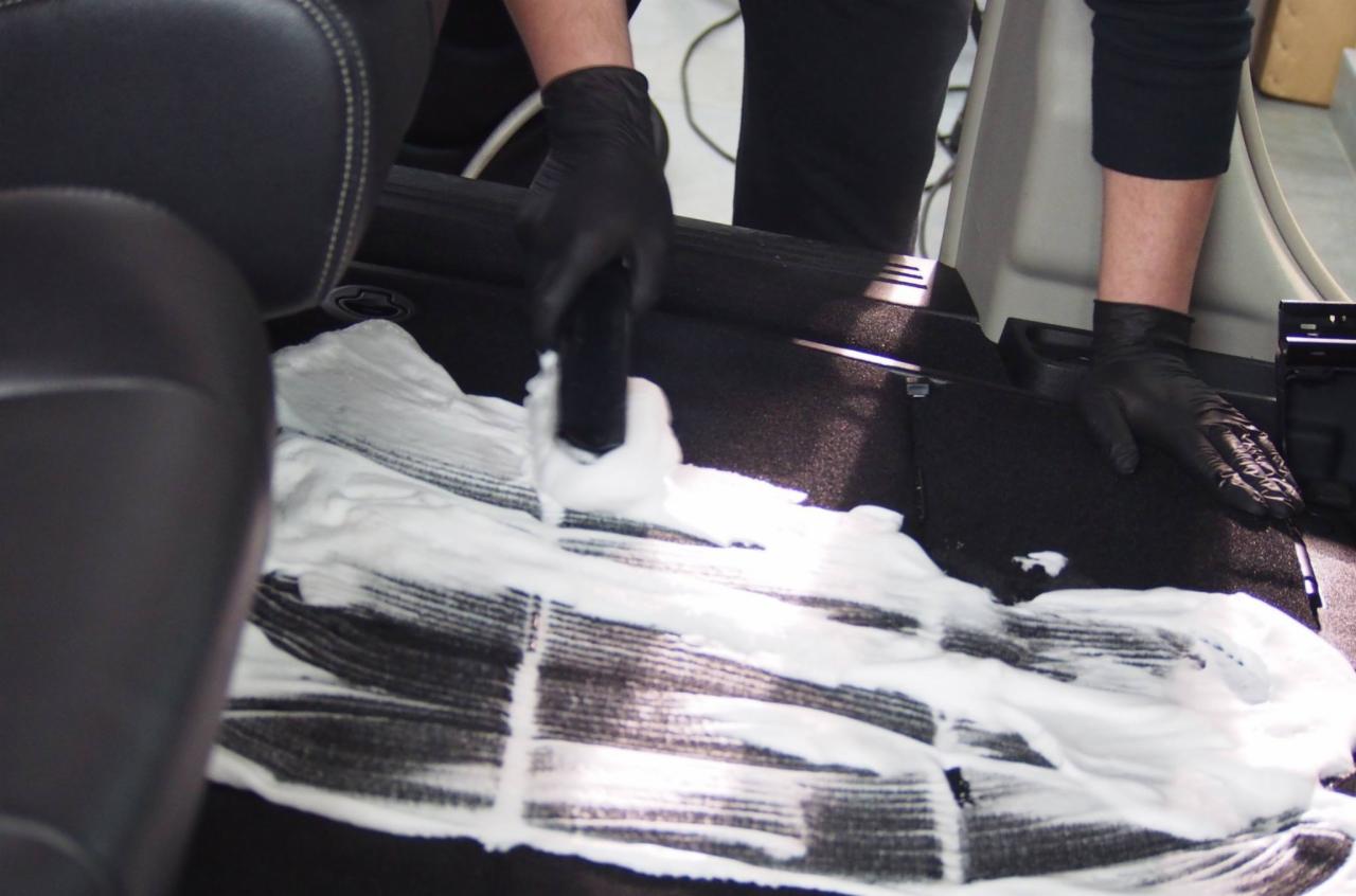 Czyszczenie tapicerki podłogowej w samochodzie za pomocą szczotki i piany, widoczne czarne rękawice ochronne.