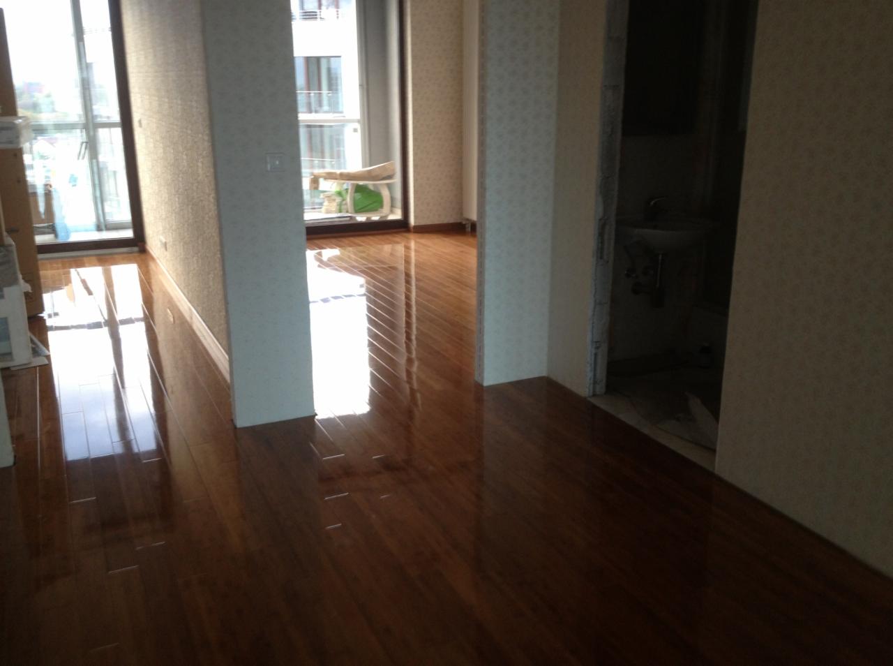 Błyszcząca, nowa podłoga z ciemnego drewna w jasnym wnętrzu mieszkania, widoczne okno balkonowe i fragment łazienki w głębi.