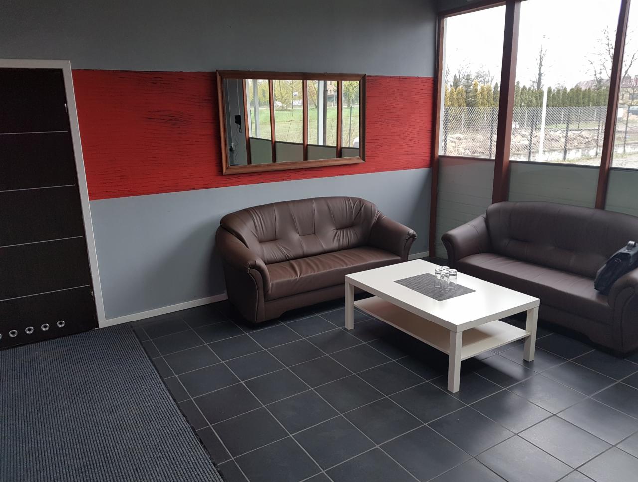 Poczekalnia z dwoma brązowymi skórzanymi sofami i białym stolikiem kawowym na czarnej podłodze z płytek. Na stoliku szklanki. Ściany w kolorze szarym i czerwonym z drewnianym lustrem. Widok...