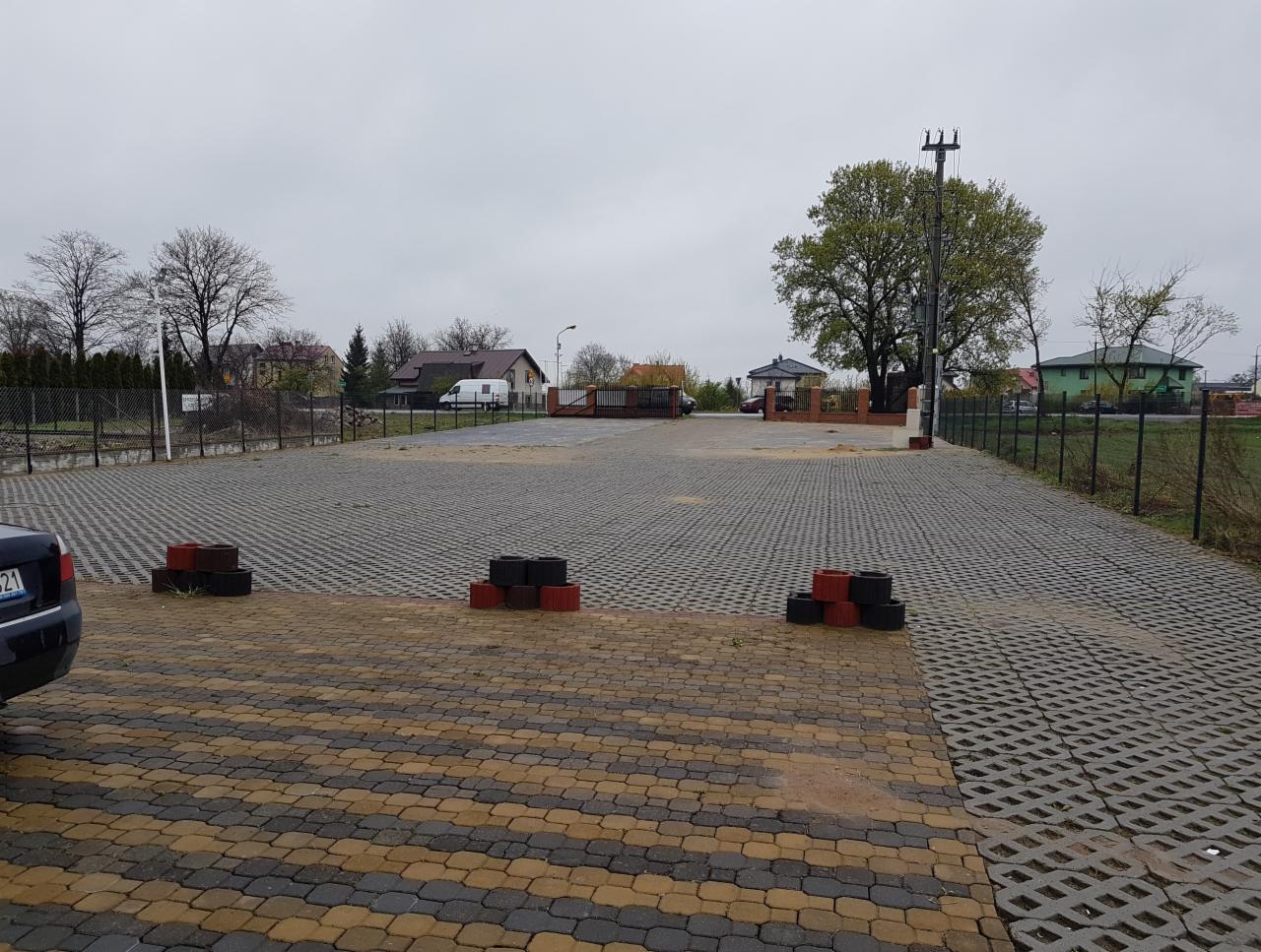 Parking z szarej ażurowej kostki, z trzema stertami czerwono-czarnych obrzeży, widok na budynki i drzewa w tle, pochmurny dzień.