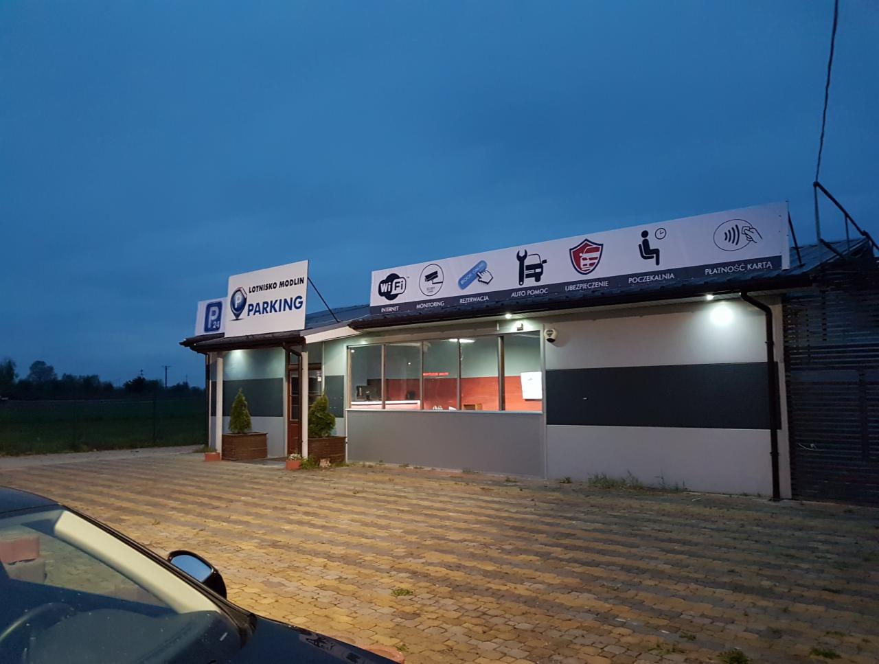 Budynek parkingu Lotnisko Modlin P24 z widocznymi oznaczeniami usług: Wi-Fi, monitoring, rezerwacja, pomoc drogowa, ubezpieczenie, poczekalnia, płatność kartą. Częściowo widoczny samochód...