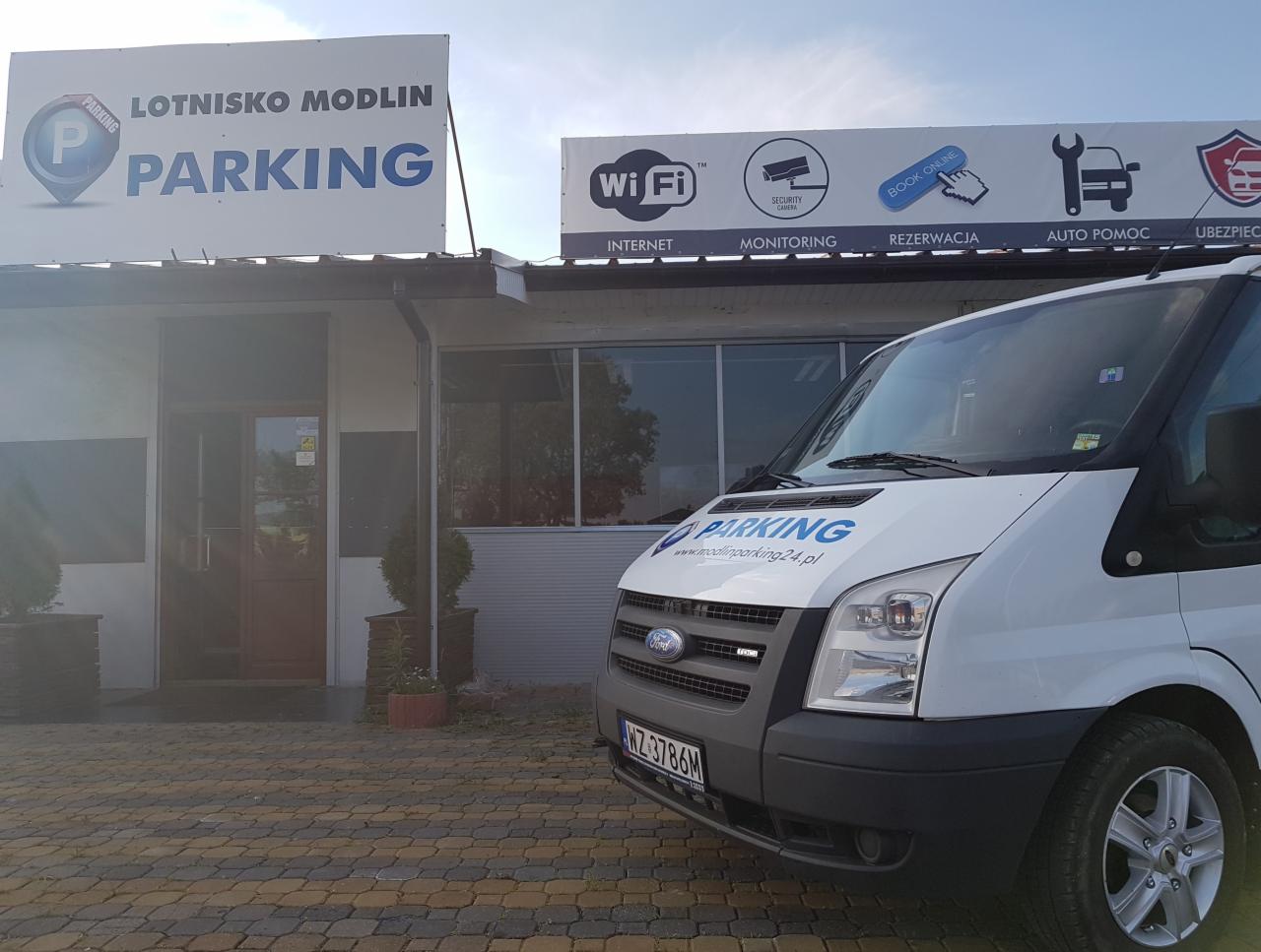 Biały bus z logo parkingu Lotnisko Modlin na tle budynku z tablicami informacyjnymi o parkingu, WiFi, monitoringu, rezerwacji online, auto pomocy i ubezpieczeniach.