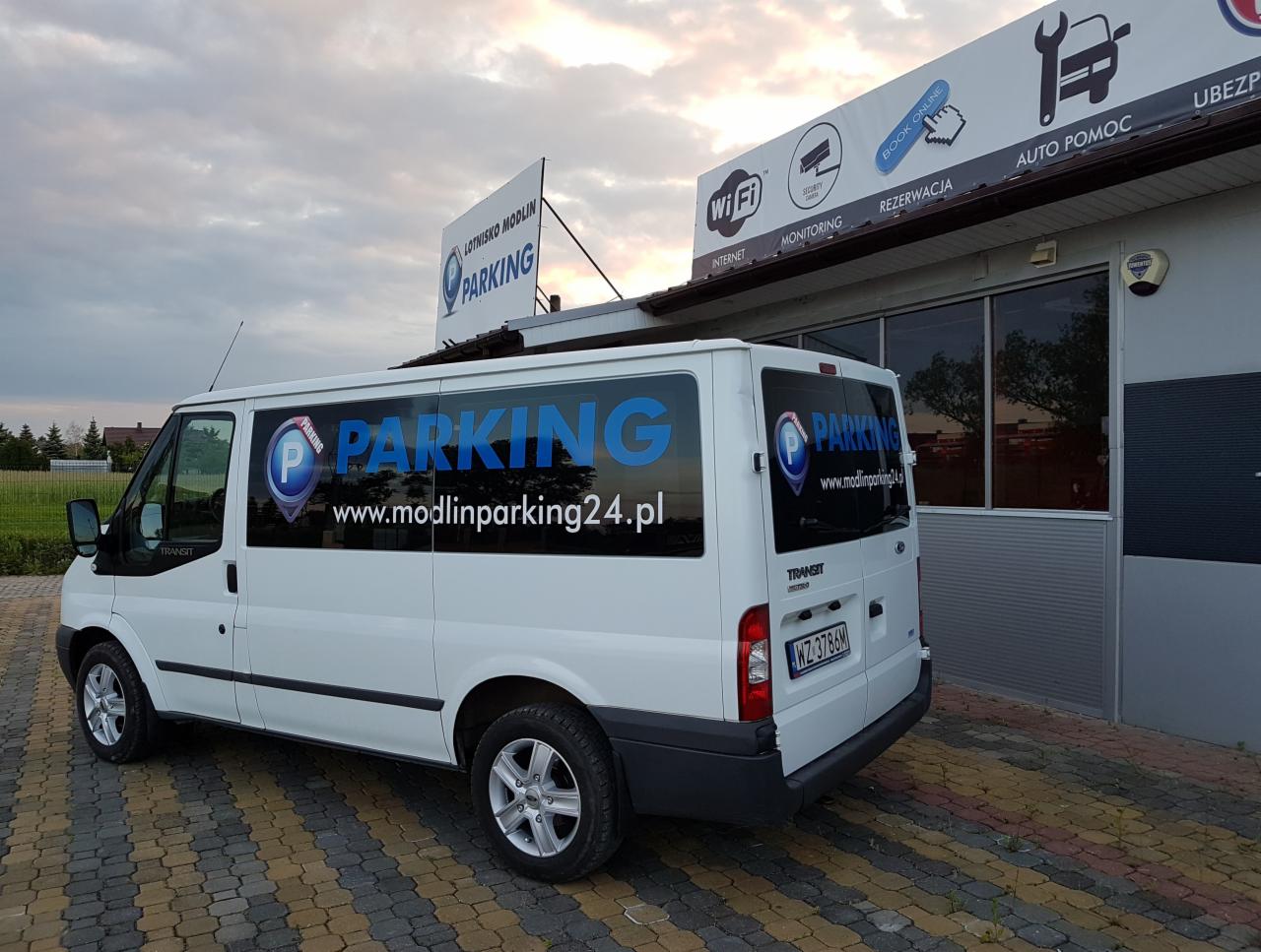 Biały bus Ford Transit z niebieskim logo parkingu Lotnisko Modlin (www.modlinparking24.pl) zaparkowany na kostce, widoczny budynek z napisem 'Auto Pomoc' i znakiem klucza francuskiego.