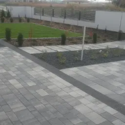Ułożona szara kostka brukowa w różnych odcieniach szarości, oddzielona pasem drobnego szarego kamienia, z widocznym trawnikiem, ogrodzeniem i logo firmy 'brukarstwo-dunajko'.