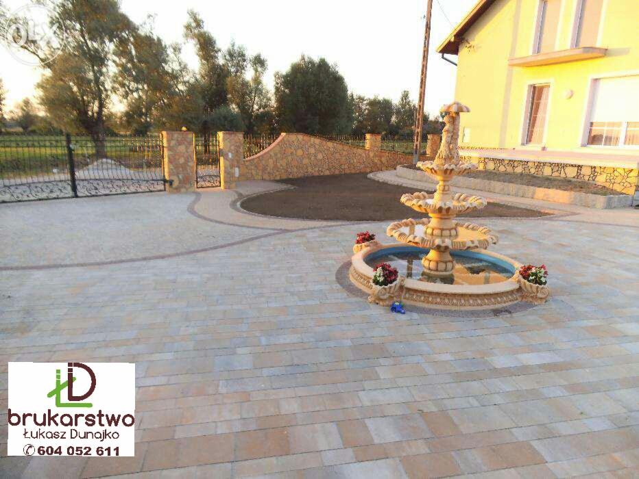 Rozległy podjazd wyłożony beżowo-szarą kostką brukową, z ozdobną, trójpoziomową fontanną po prawej stronie, ogrodzeniem z kutego żelaza i murem z kamienia w tle oraz logo firmy w lewym dolnym rogu.