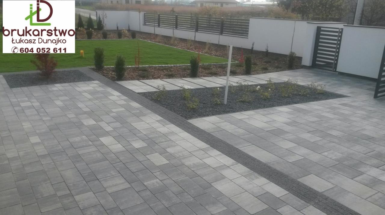 Ułożona szara kostka brukowa w różnych odcieniach szarości, oddzielona pasem drobnego szarego kamienia, z widocznym trawnikiem, ogrodzeniem i logo firmy 'brukarstwo-dunajko'.