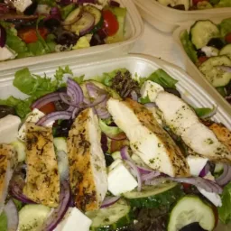 Sałatka grecka z grillowanym kurczakiem w plastikowym pojemniku, widoczne liście sałaty, ogórek, pomidory, czerwona cebula, oliwki i ser feta.