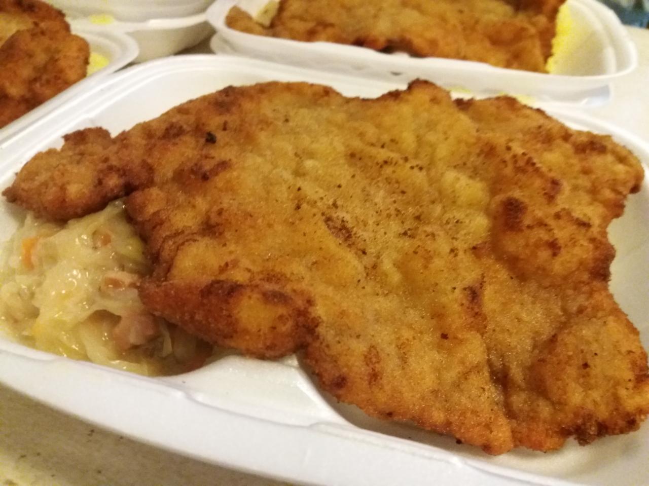 Duży, panierowany sznycel wieprzowy z kapustą duszoną w białym, styropianowym pojemniku na wynos.