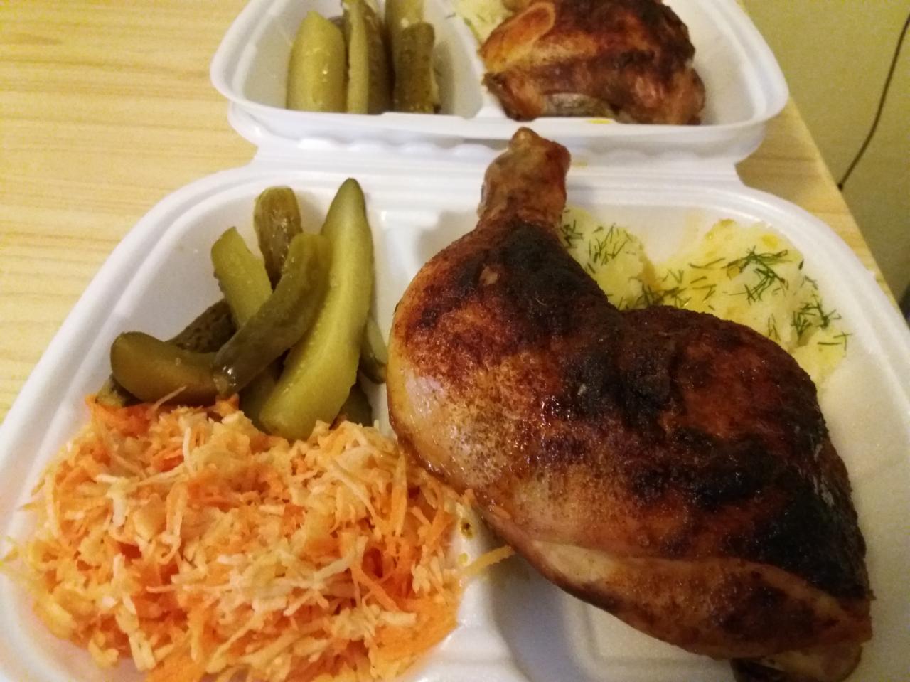 Pieczone udo kurczaka z ziemniakami z koperkiem, surówką z marchewki i ogórkami kiszonymi w białym styropianowym pojemniku na wynos.