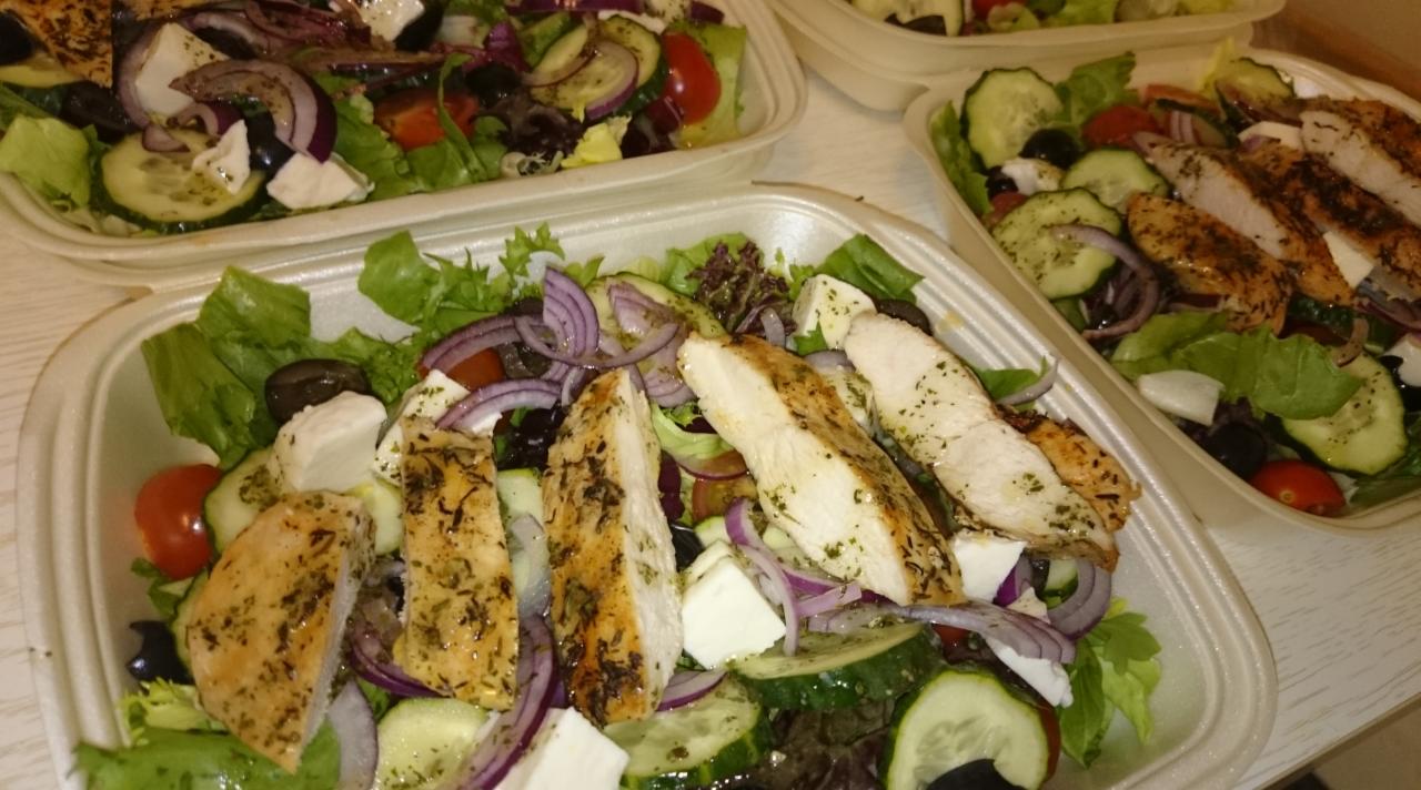 Sałatka grecka z grillowanym kurczakiem w plastikowym pojemniku, widoczne liście sałaty, ogórek, pomidory, czerwona cebula, oliwki i ser feta.