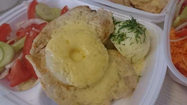 Gotowe danie obiadowe w białym plastikowym pojemniku: pierś z kurczaka polana sosem serowym i ananasem, ziemniaki z koperkiem oraz sałatka warzywna z pomidorów, ogórków i cebuli.