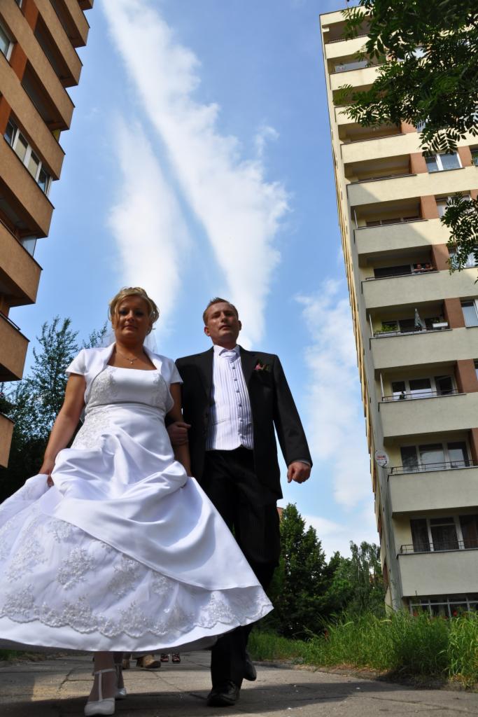 Para młoda, panna młoda w białej sukni z koronkowymi zdobieniami i pan młody w czarnym garniturze, spacerują po chodniku na tle bloków mieszkalnych i błękitnego nieba z chmurami.