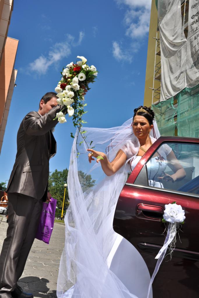 Panna młoda w sukni ślubnej wychodząca z bordowego samochodu udekorowanego białą różą, mężczyzna w garniturze trzyma bukiet kwiatów na tle błękitnego nieba i budynku w rusztowaniach.