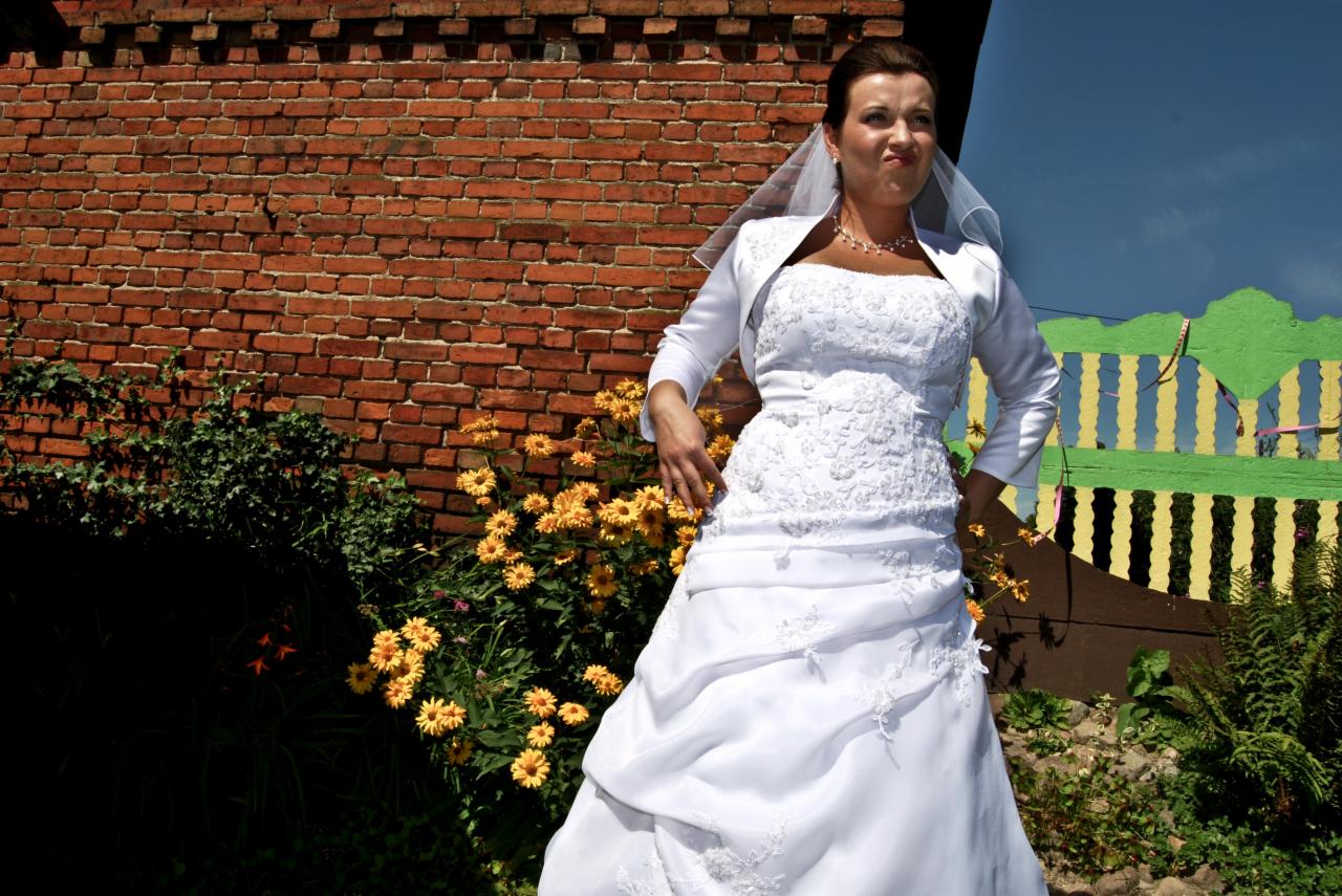 Panna młoda w białej sukni ślubnej i bolerku, pozująca na tle ceglanej ściany i kolorowego płotu, z kwiatami w ogrodzie, z grymasem na twarzy.
