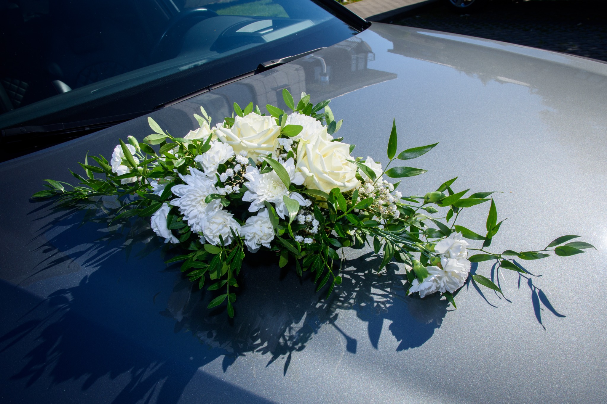 Dekoracja z białych róż, chryzantem i zielonych liści na srebrnym samochodzie, widok z góry, ostre światło słoneczne.